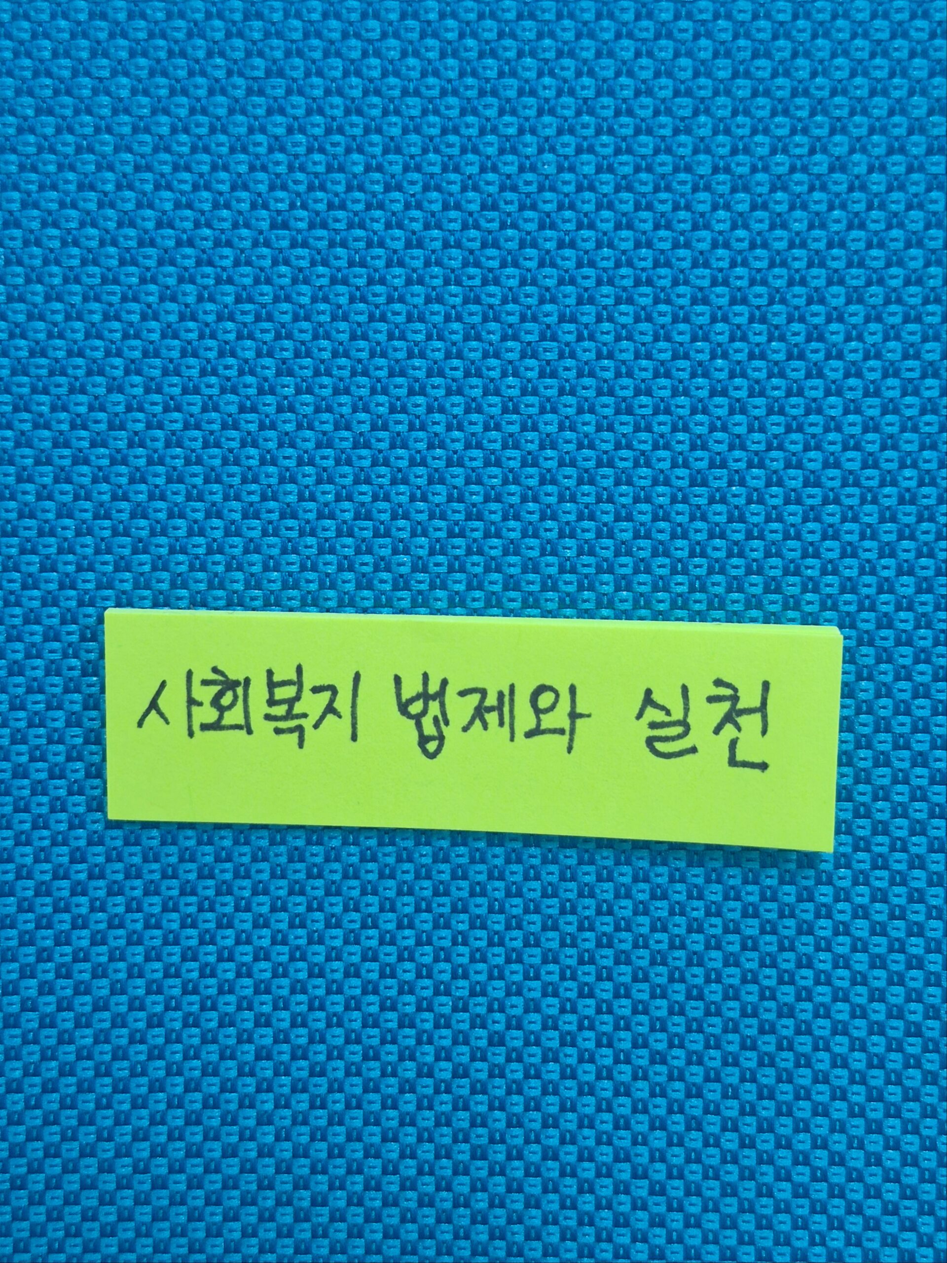 '사회복지 법제와 실천'이라고 손글씨로 적힌 노란색 메모지가 파란색 직물 질감의 배경에 붙어 있는 모습