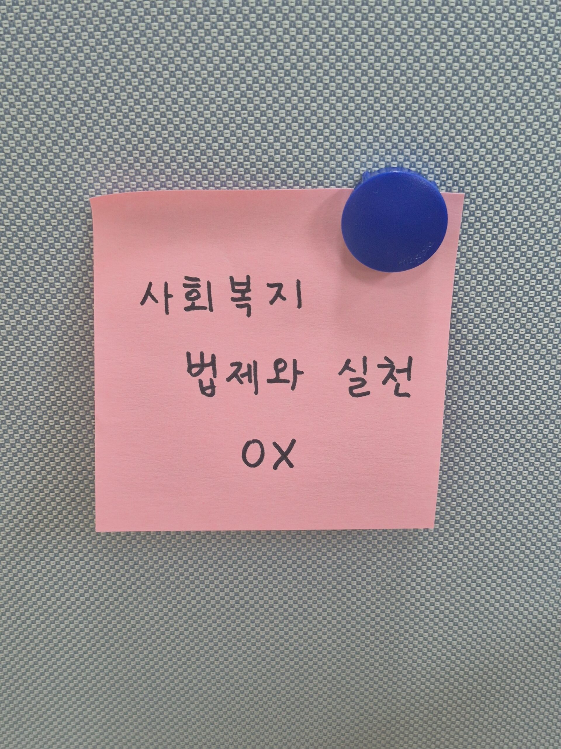 회색 파티션에 파란색 원형 자석으로 고정된 핑크색 포스트잇 사진. 포스트잇에는 검은색 손글씨로 '사회복지 법제와 실천 OX'라고 적혀 있다.