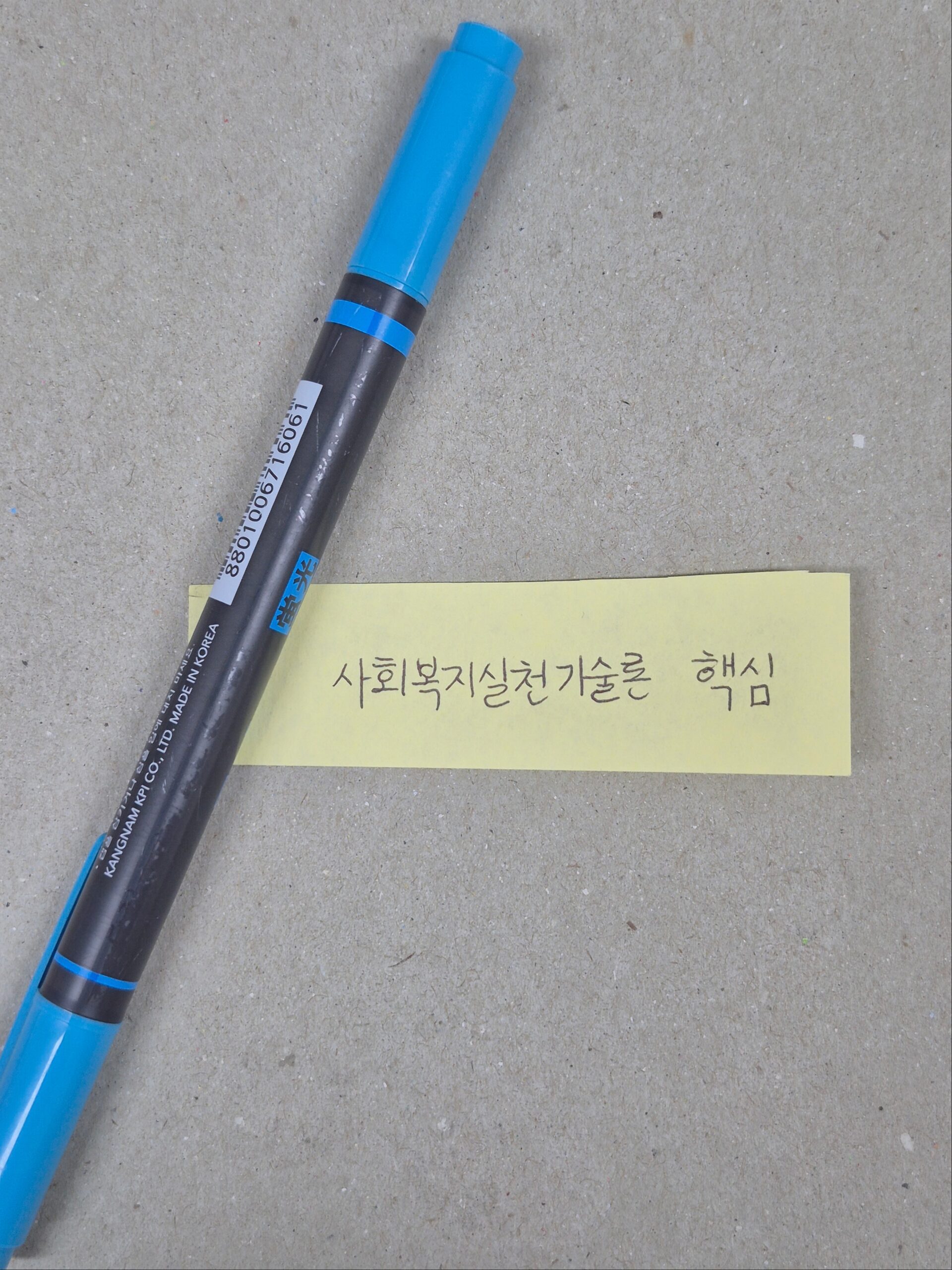 회색 바탕 위에 파란색 형펜이 놓여 있고, 그 아래에 "사회복지실천기술론 핵심"이라고 한글로 손글씨가 적힌 노란색 포스트잇이 붙어 있는 사진.