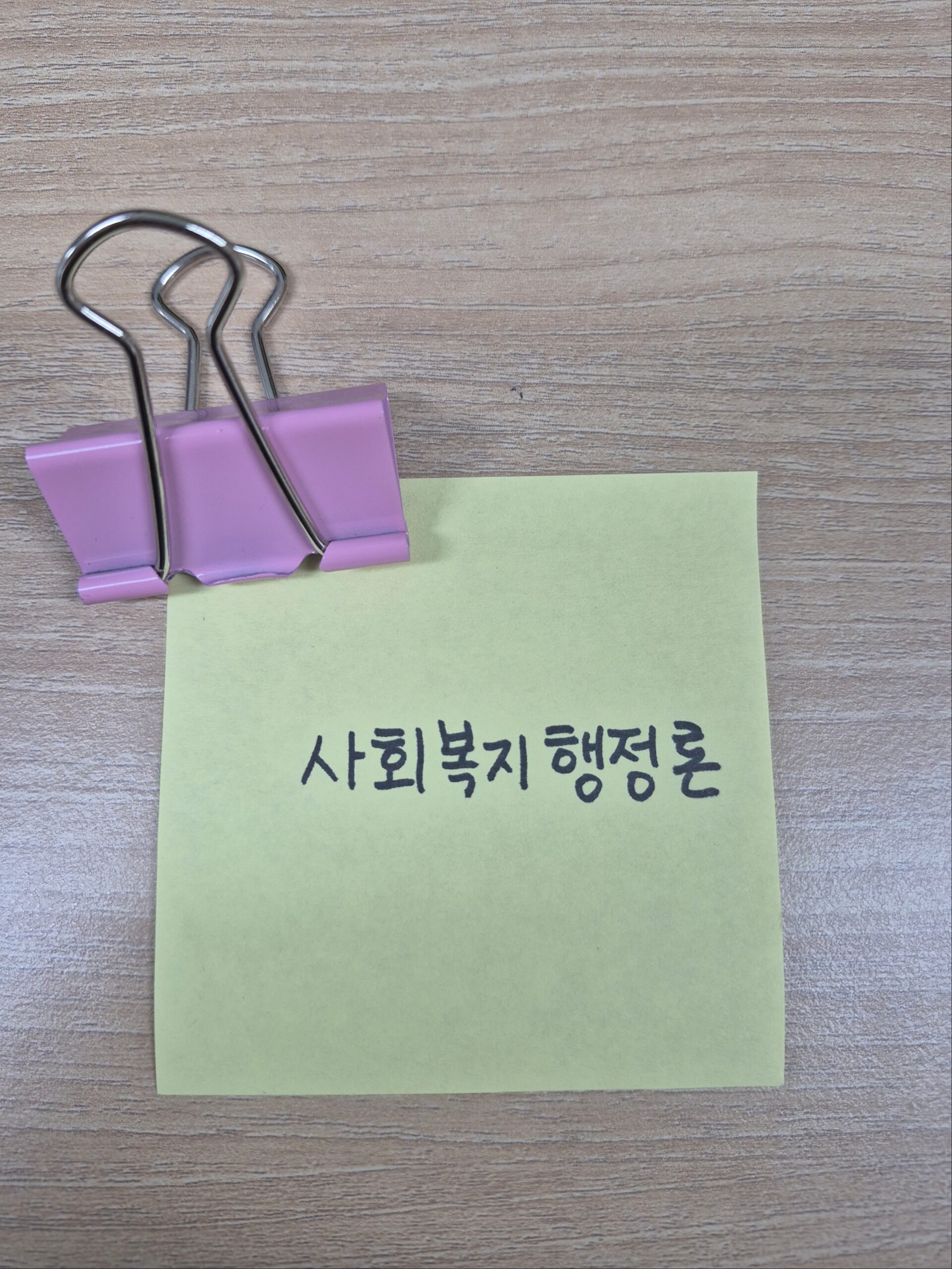 나뭇결 책상 위에 분홍색 바인더 클립으로 고정된 연두색 포스트잇. 포스트잇에는 검은색 손글씨로 '사회복지행정론'이라고 적혀 있다. 사회복지사 1급 핵심 요약을 상징하는 대표 이미지.