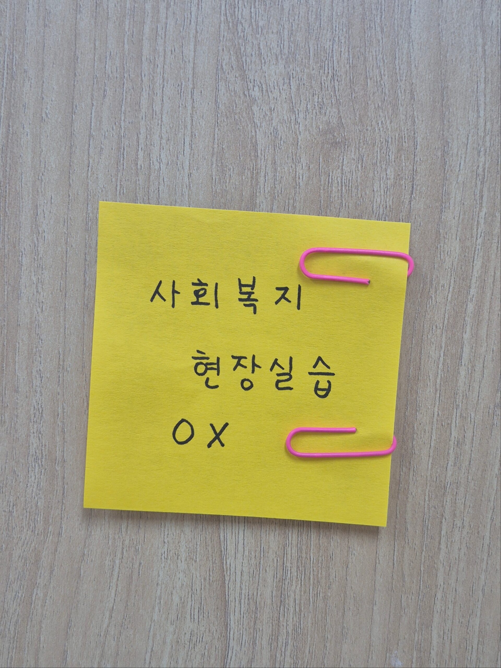 밝은 나무 테이블 위에 '사회복지 현장실습 OX'라고 손글씨로 적힌 노란색 포스트잇이 놓여 있습니다. 포스트잇의 오른쪽 상단과 하단에는 분홍색 클립 두 개가 꽂혀 있어 학습 자료나 중요한 메모임을 나타냅니다.