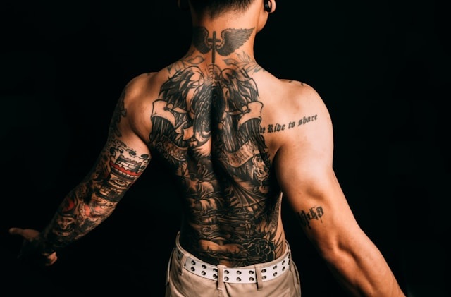 검은색 배경 앞에서 상의를 탈의한 채 등을 돌리고 있는 남성의 모습. 목 뒤의 날개와 십자가 문신을 시작으로 등 전체와 팔에 화려하고 정교한 블랙앤그레이 타투가 가득 새겨져 있다. 영화 메멘토의 문신 모티브를 연상시키는 강렬한 뒷모습 이미지.