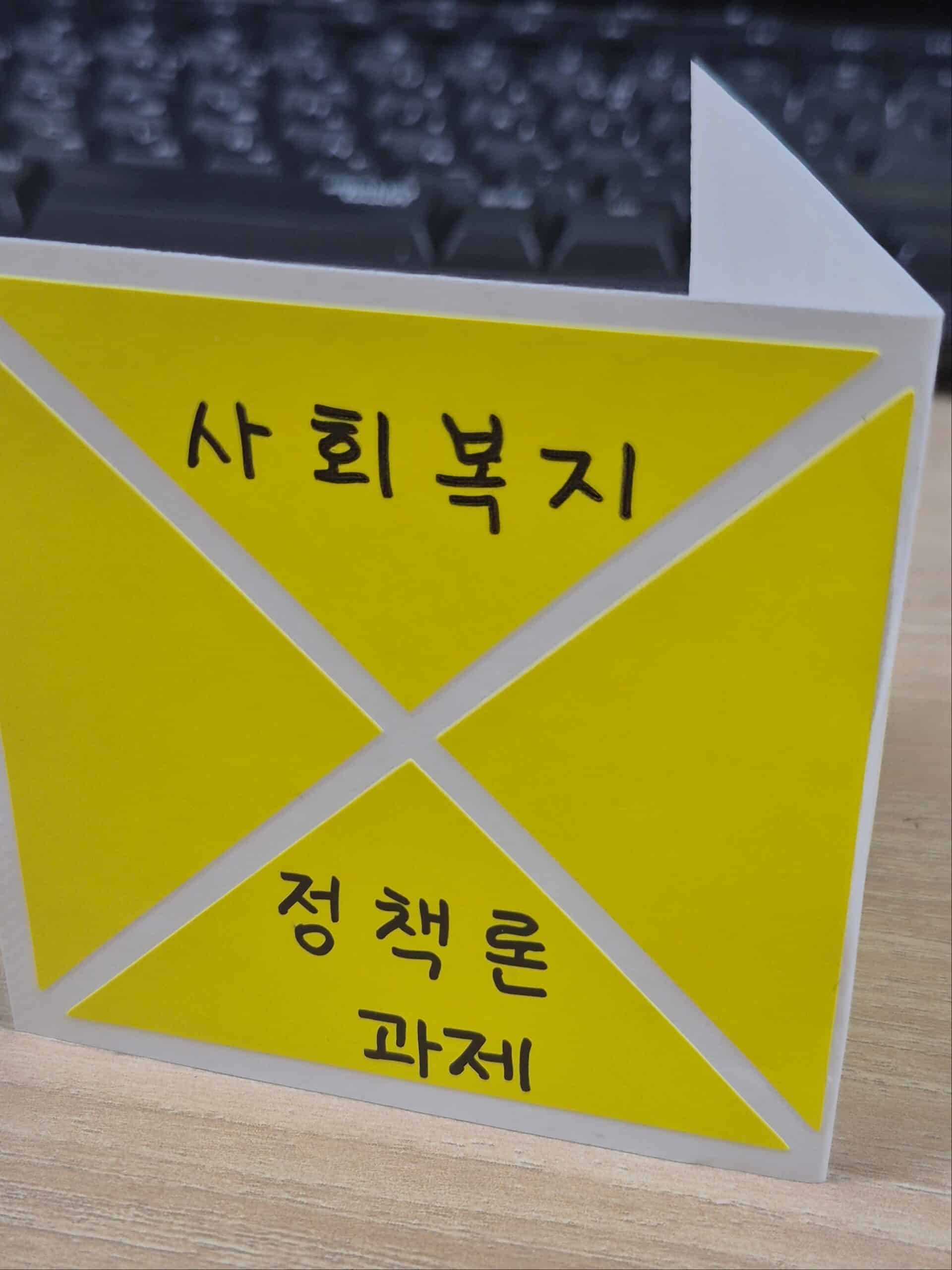 책상 위 키보드를 배경으로, '사회복지', '정책론', '과제'라고 적힌 노란색 종이가 붙어 있는 입체적인 흰색 삼각형 구조물 이미지
