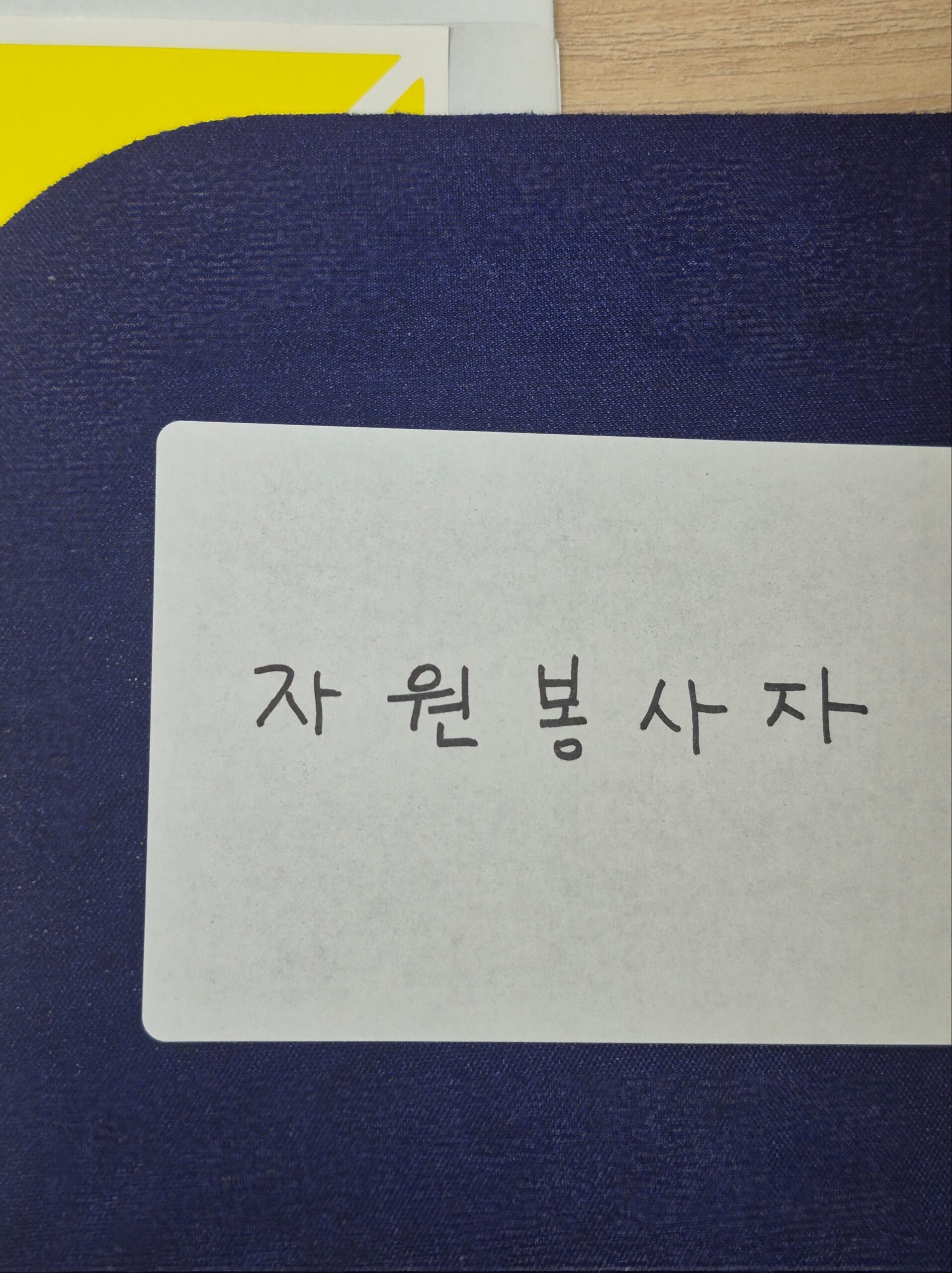 남색 바닥 위에 놓인 흰색 종이에 손글씨로 '자원봉사자'라고 적혀 있는 사진. 사회복지학 자원봉사론 과제와 실제 봉사 경험을 대표하는 이미지.