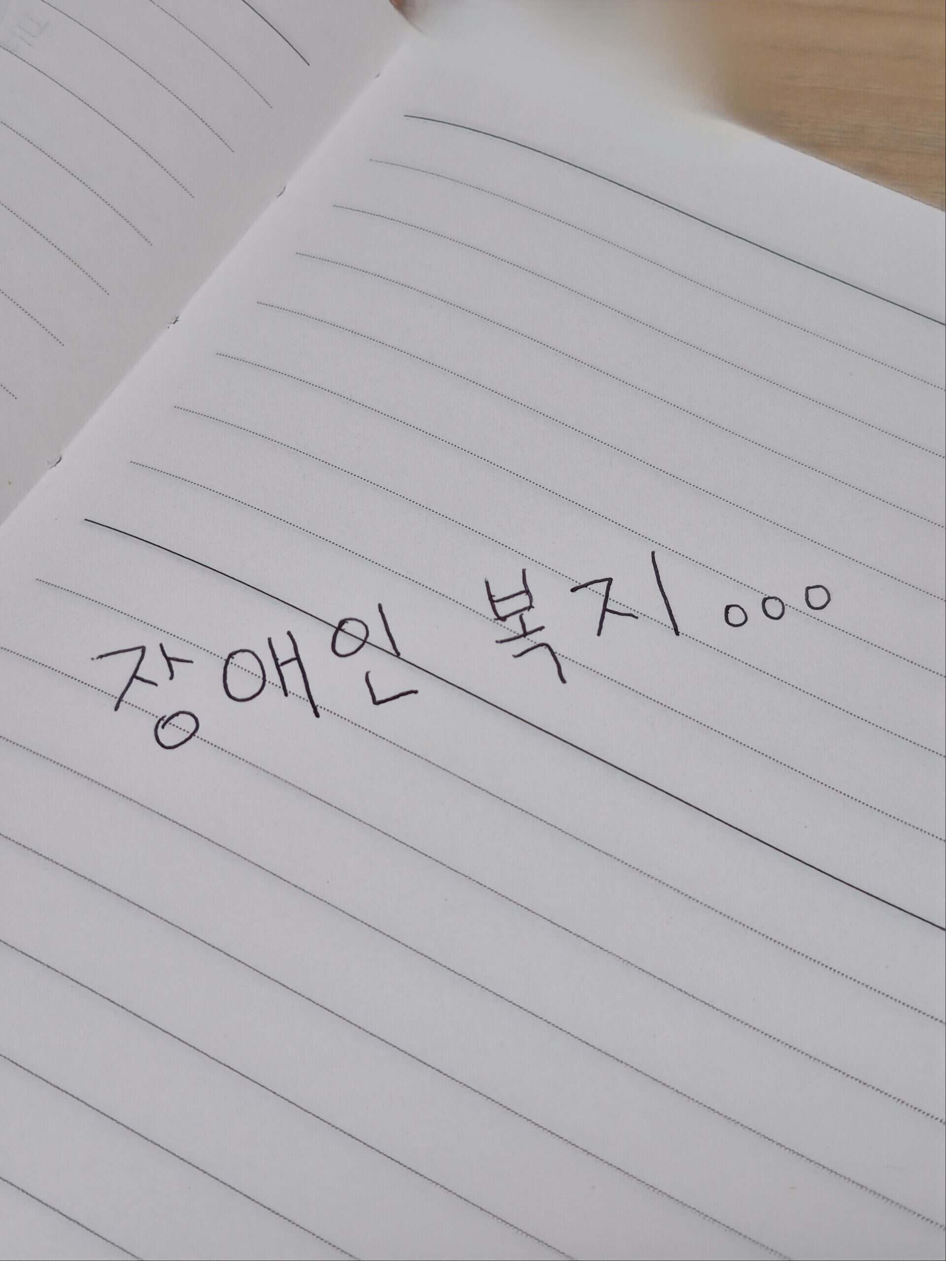 줄 그어진 공책에 검은색 펜으로 손수 쓴 '장애인 복지'라는 한글 문구.